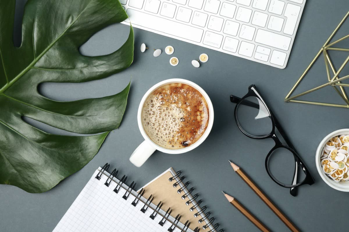 Coffee, glasses, pencils and a spiral notebook beside a keyboard on a gray desk